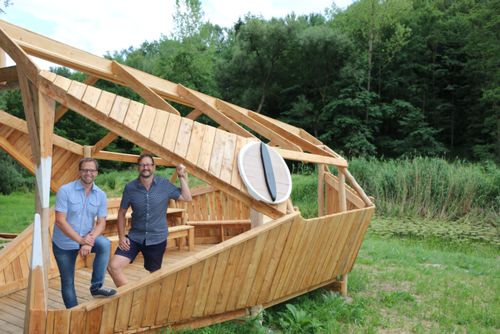 Zwei Männer in Freizeitkleidung posieren vor einer hölzernen Struktur, die einem Boot ähnelt, in einem üppigen grünen Wald.