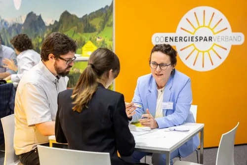 Drei Personen sitzen an einem Tisch, zwei Frauen und ein Mann. Die Frau im blauen Blazer spricht zu den anderen beiden. Sie sind bei einer Veranstaltung mit einem gelben Hintergrund und einem Logo.
