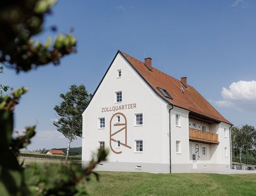 Ein weißes Gebäude mit rotem Dach namens Zollquartier, umgeben von einem grünen Rasen und Bäumen.