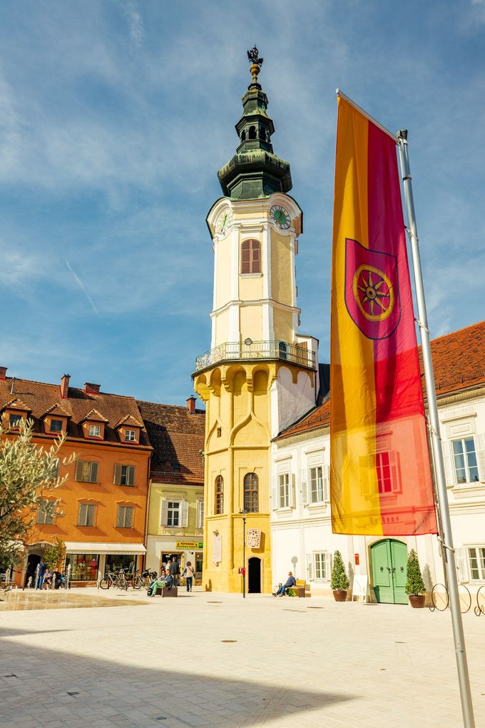  Abschnitt Bild: Ein gelber Turm mit einer Uhr steht vor einem weißen Gebäude. Eine rot-gelbe Flagge befindet sich rechts neben dem Turm.