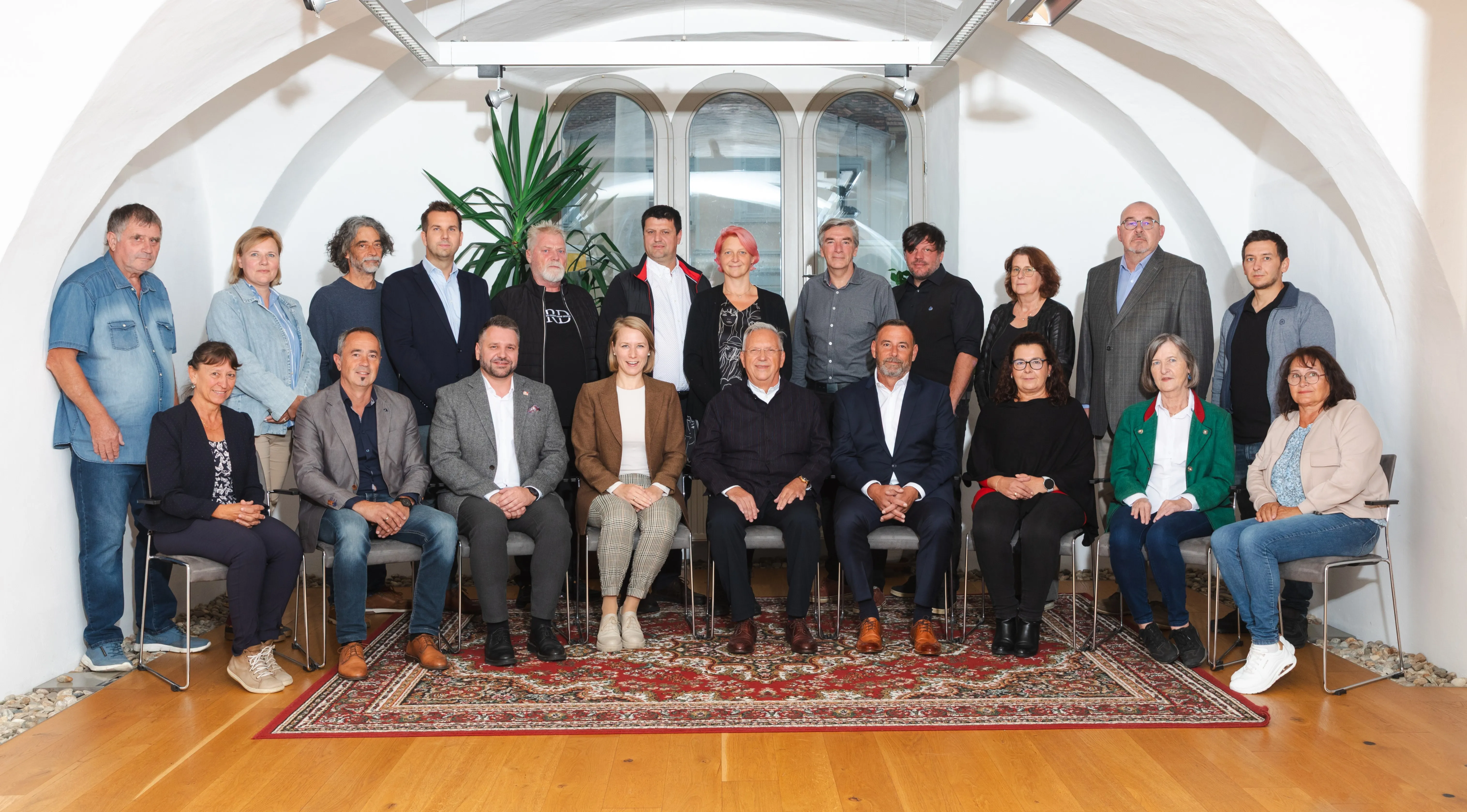 Eine Gruppe von Menschen, sowohl Männer als auch Frauen, sitzen im Halbkreis auf Stühlen in einem Raum. Sie schauen alle in die gleiche Richtung, wahrscheinlich für ein Foto. Hinter ihnen steht eine große Pflanze und ein Fenster.