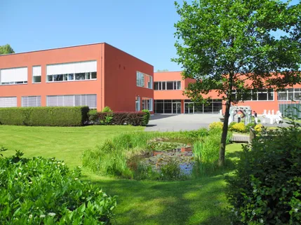 Ein rotes Gebäude mit Glasfenstern befindet sich in einer grünen Landschaft. Das Gebäude hat einen großen Vorgarten mit Rasen, Büschen und einem kleinen Teich mit Seerosen. Ein Baum steht in der Nähe des Teichs, und eine Skulptur befindet sich im Hintergrund.
