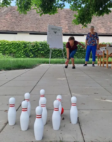 Zwei ältere Frauen spielen eine Partie Bowling auf einem Betonplatz. Eine Frau beugt sich vor, um den Ball zu werfen, während die andere neben einem Whiteboard steht.