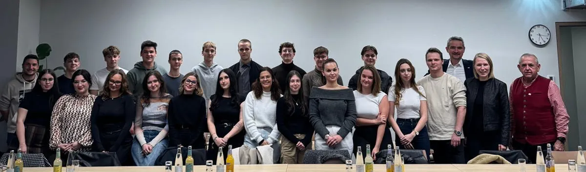 Eine Gruppe von Personen steht und sitzt gemeinsam in einem Konferenzraum, lächelt und posiert für ein Foto. Vor ihnen sind Flaschen mit Getränken auf einem Tisch angeordnet.