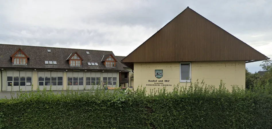 Ein Gebäude mit braunem Dach und einem Logo an der Seite, mit einem grünen Busch davor und einem weiteren Gebäude mit Glasfenstern und Solarpanelen.