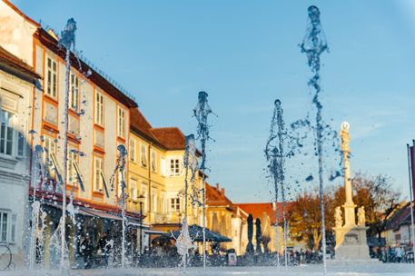 Wasserbrunnen auf einem Stadtplatz mit Menschen und Gebäuden. Ein blauer Himmel darüber, mit Bäumen und einem Denkmal sichtbar.