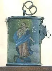 Eine kleine, antike Blechdose, bemalt mit einem Bild der Jungfrau Maria, die ein Baby Jesus hält. Die Dose hat oben einen Metallgriff und steht auf einer Holzoberfläche.