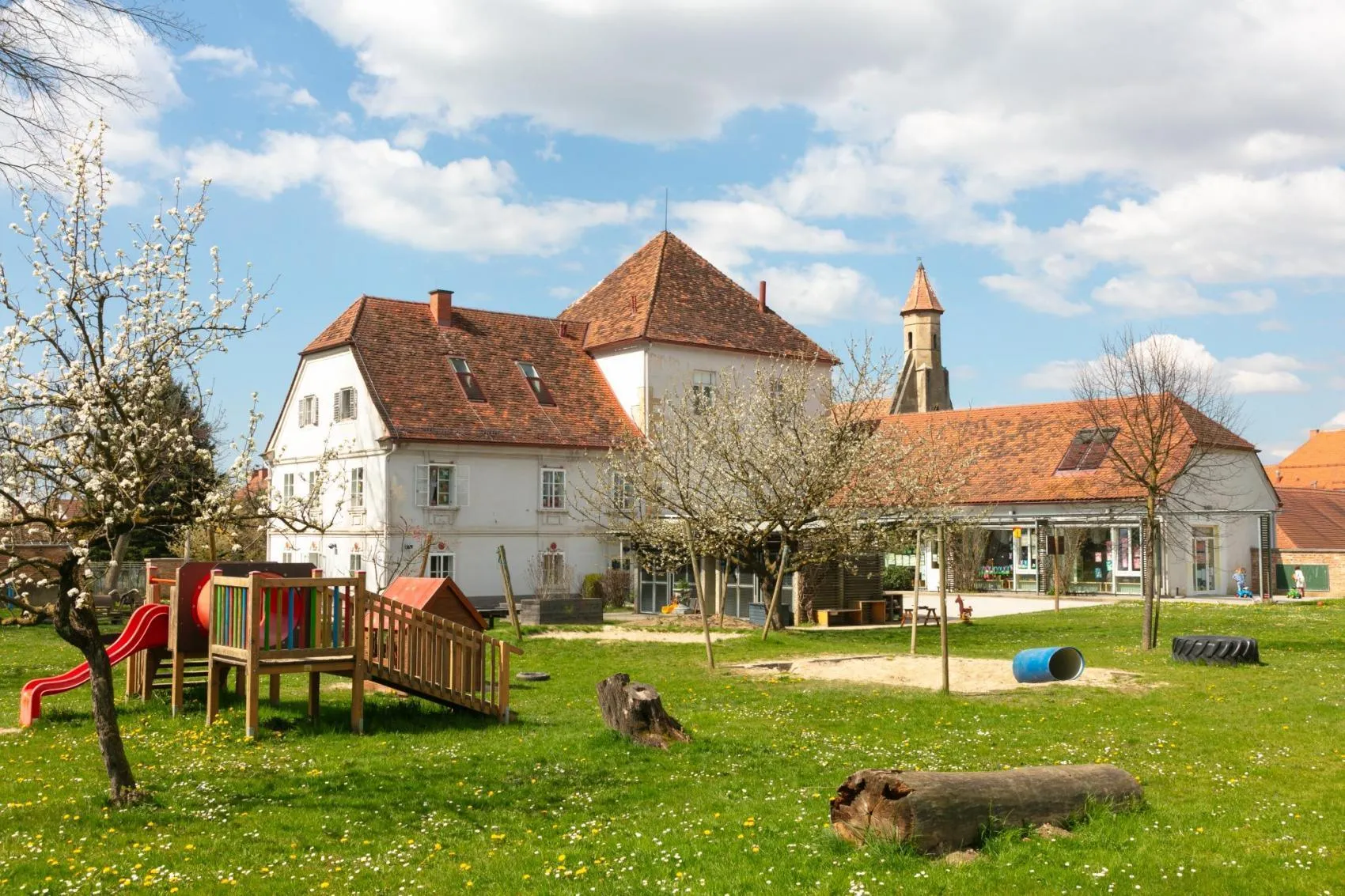 Ein großes weißes Gebäude mit braunen Dächern steht in einem grünen Hof. In der Nähe befindet sich ein Spielplatz mit Rutsche und einem kleinen Haus. Bäume und Blumen sind in der Umgebung.