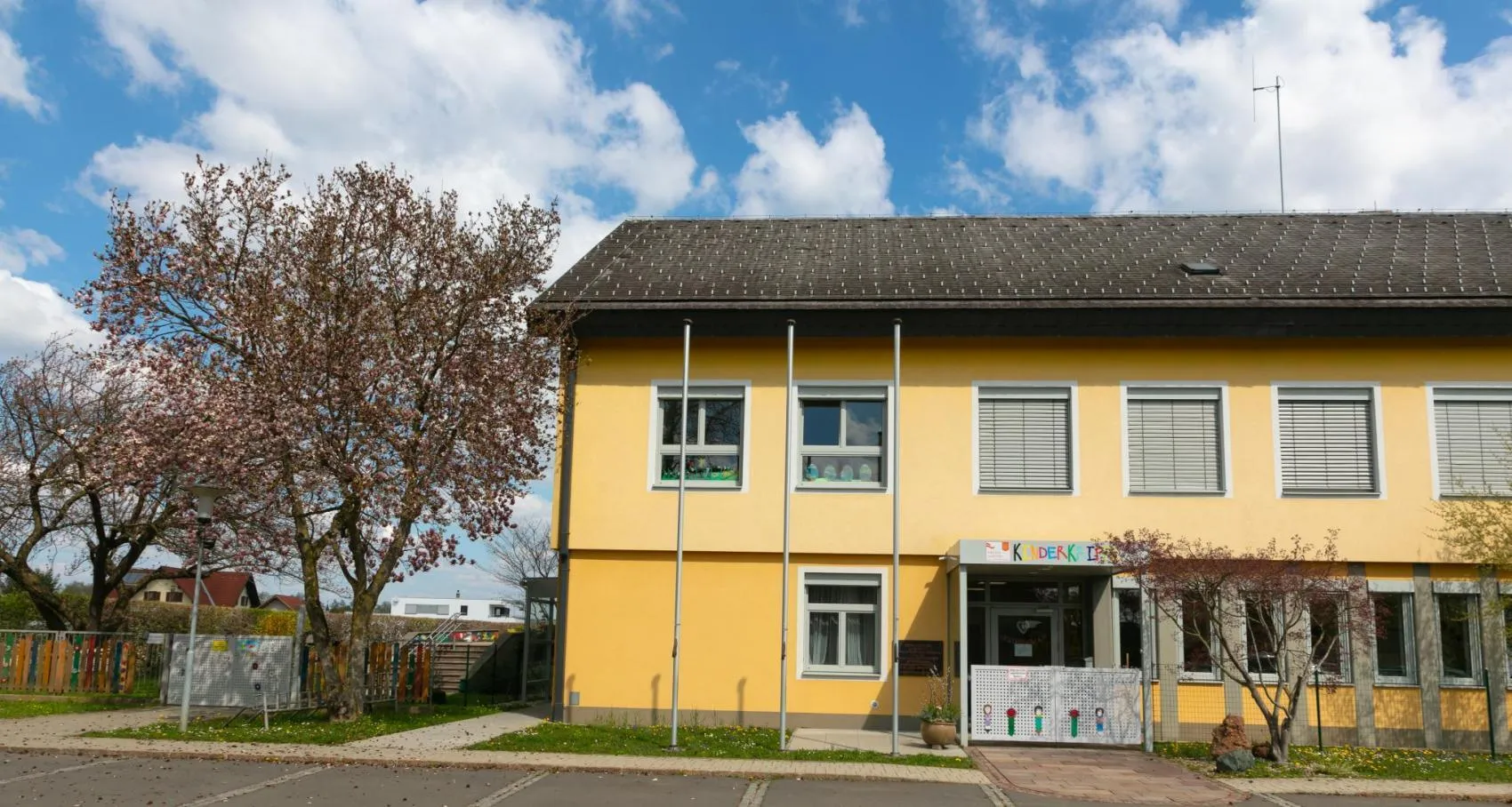 Ein leuchtend gelbes Gebäude mit mehreren Fenstern und einem Schild über dem Eingang, das 'Kinderkip' steht. Das Gebäude hat ein schwarzes Dach und ist von einem Baum und einem kleinen Rasenbereich umgeben.