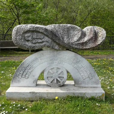 Eine große Steinskulptur in einem Graspark mit einer Bank, Bäumen und Blumen. Die Skulptur zeigt ein abstraktes Design mit einem Rad und Text.