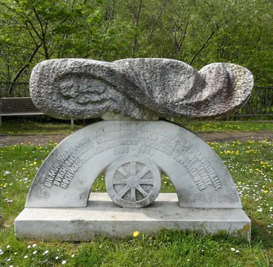 Eine Steinskulptur mit einem Bogen und einem Rad steht in einem Grasbereich. Bäume und eine Bank sind im Hintergrund. Die Skulptur hat einen gebogenen Bogen und ein Rad an ihrer Basis.