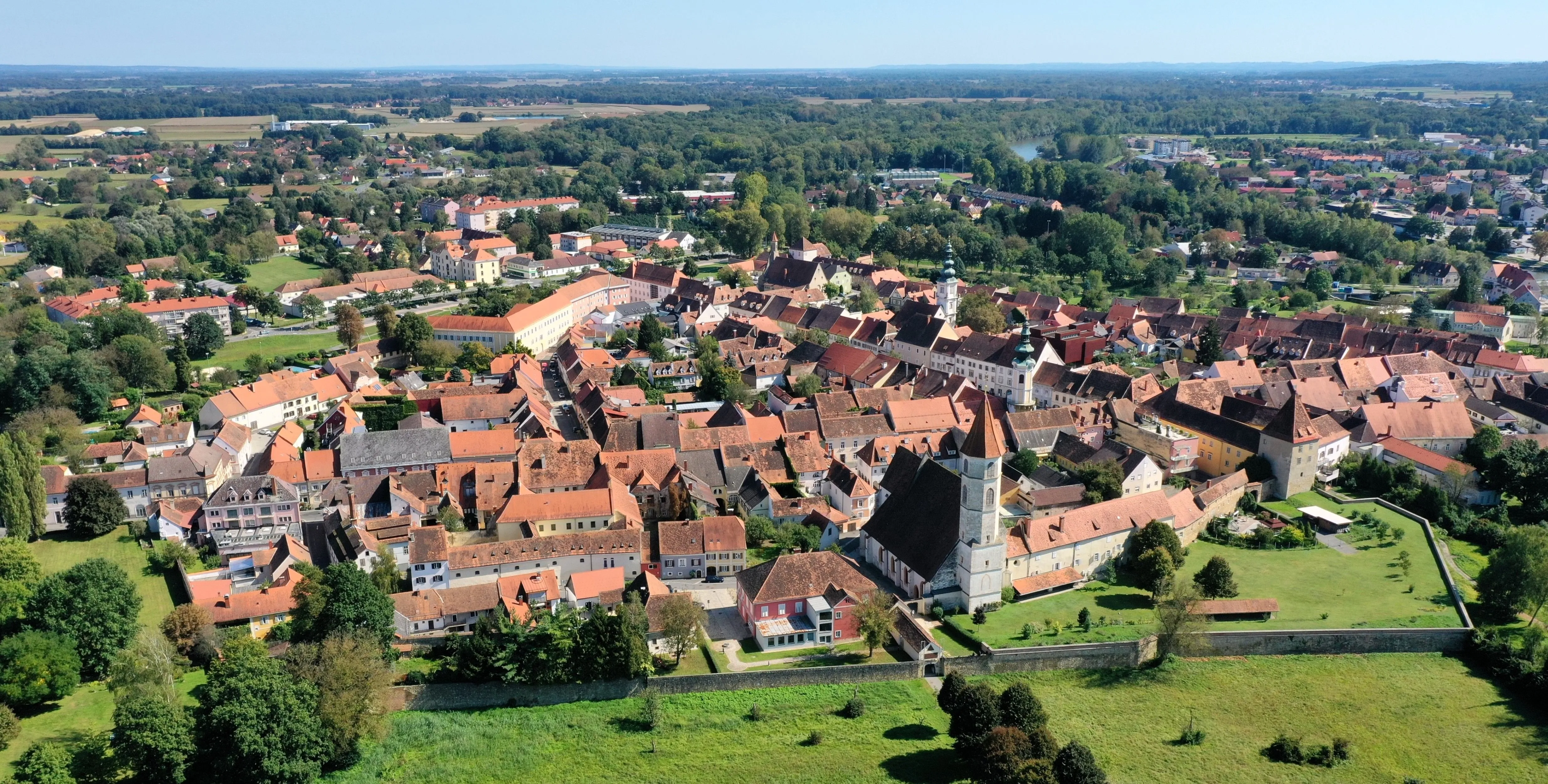 Luftaufnahme einer historischen Stadt mit zahlreichen Gebäuden, meist mit roten Dächern. Ein markanter Kirchturm ist sichtbar, umgeben von üppigem Grün und Bäumen. Die Stadt liegt in einem Tal mit einem Fluss und Hügeln im Hintergrund.