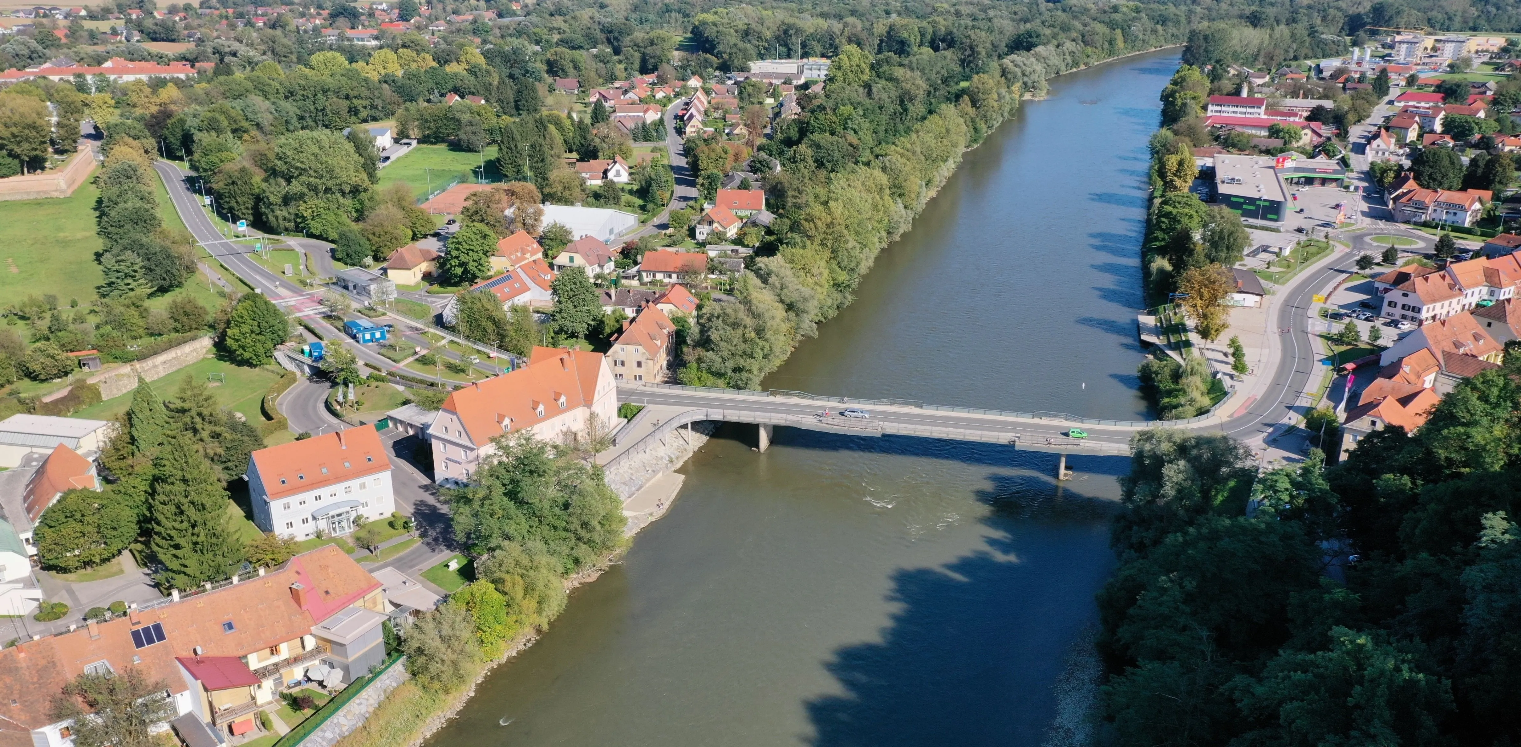 Eine Luftaufnahme einer Stadt mit Häusern und einer Brücke über einem Fluss. Autos fahren auf der Brücke, und die Umgebung ist von Bäumen und viel Grün geprägt.