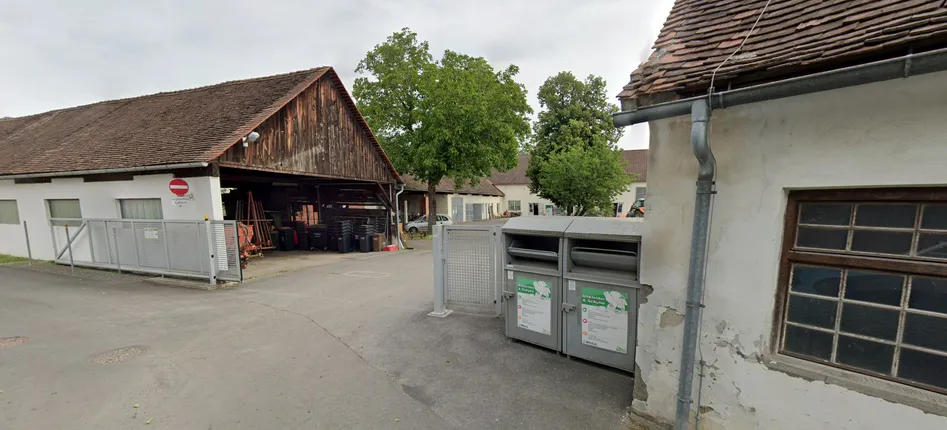 Ein Hof mit zwei grauen Containern für Kleidungsspenden. Ein Scheune und mehrere Bäume im Hintergrund.