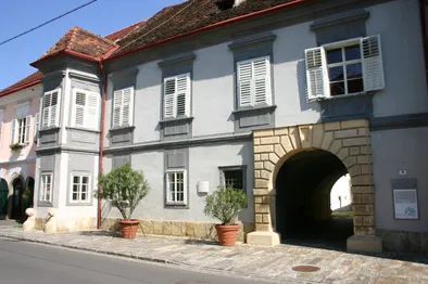 Ein weißes Gebäude mit rotem Rand und Fensterläden, zwei Topfpflanzen auf dem Bürgersteig, ein Steinbogeneingang und ein rotes Dach.