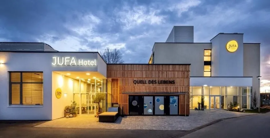 Der Eingangsbereich eines Hotels mit dem Namen JUFA Hotel an der Wand. Es gibt Pflanzen und Bäume an der Seite. Der Eingang hat Glastüren mit runden Fenstern.