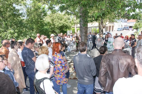 Eine große Gruppe von Menschen versammelt sich im Freien, möglicherweise bei einer Veranstaltung. Einige stehen, andere gehen. Bäume und ein Gebäude sind im Hintergrund zu sehen.