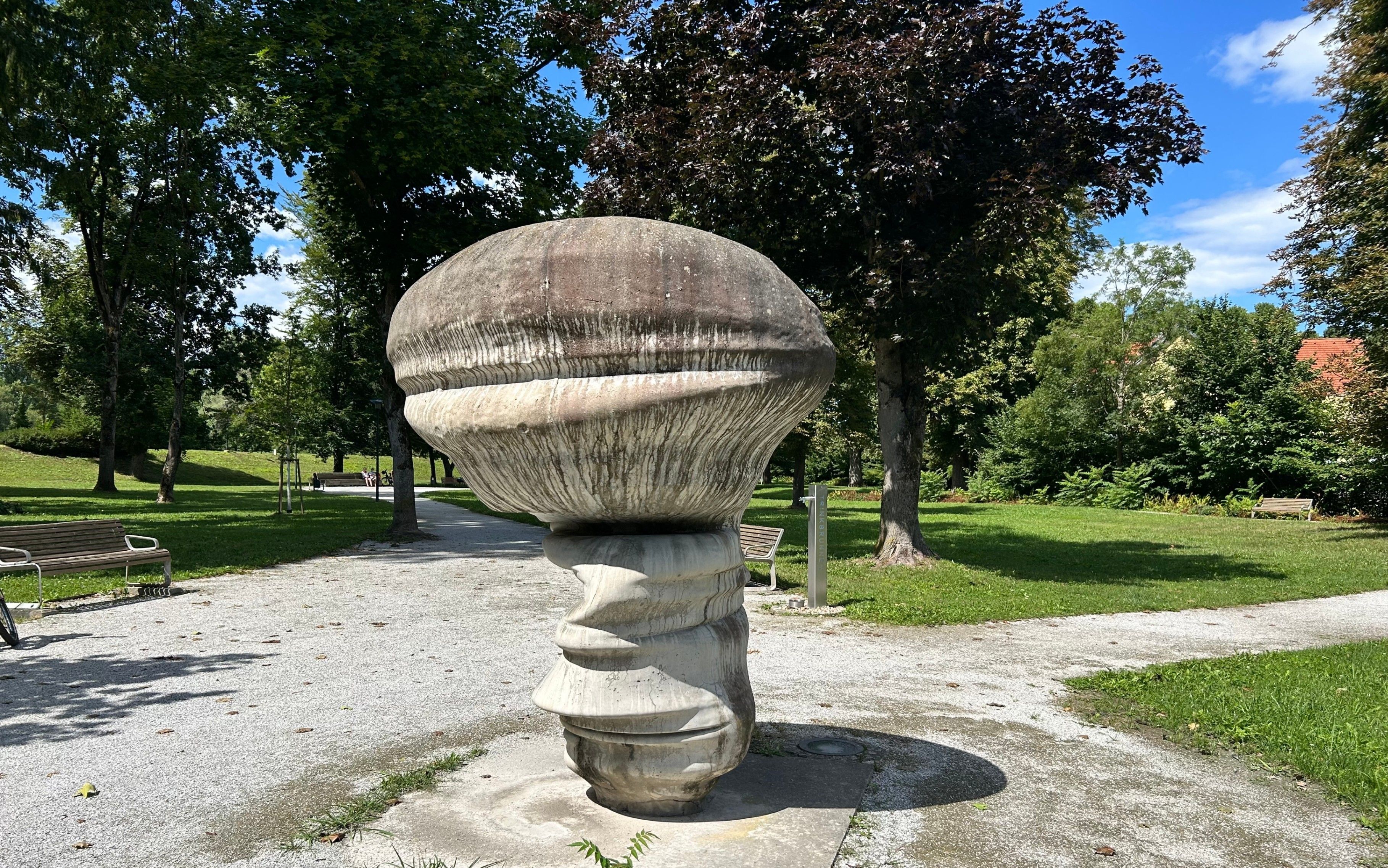 Eine große Steinskulptur, die einem Pilz ähnelt, steht auf einem Betonsockel in einem Park. Sie ist von saftigem grünem Gras und Bäumen umgeben.