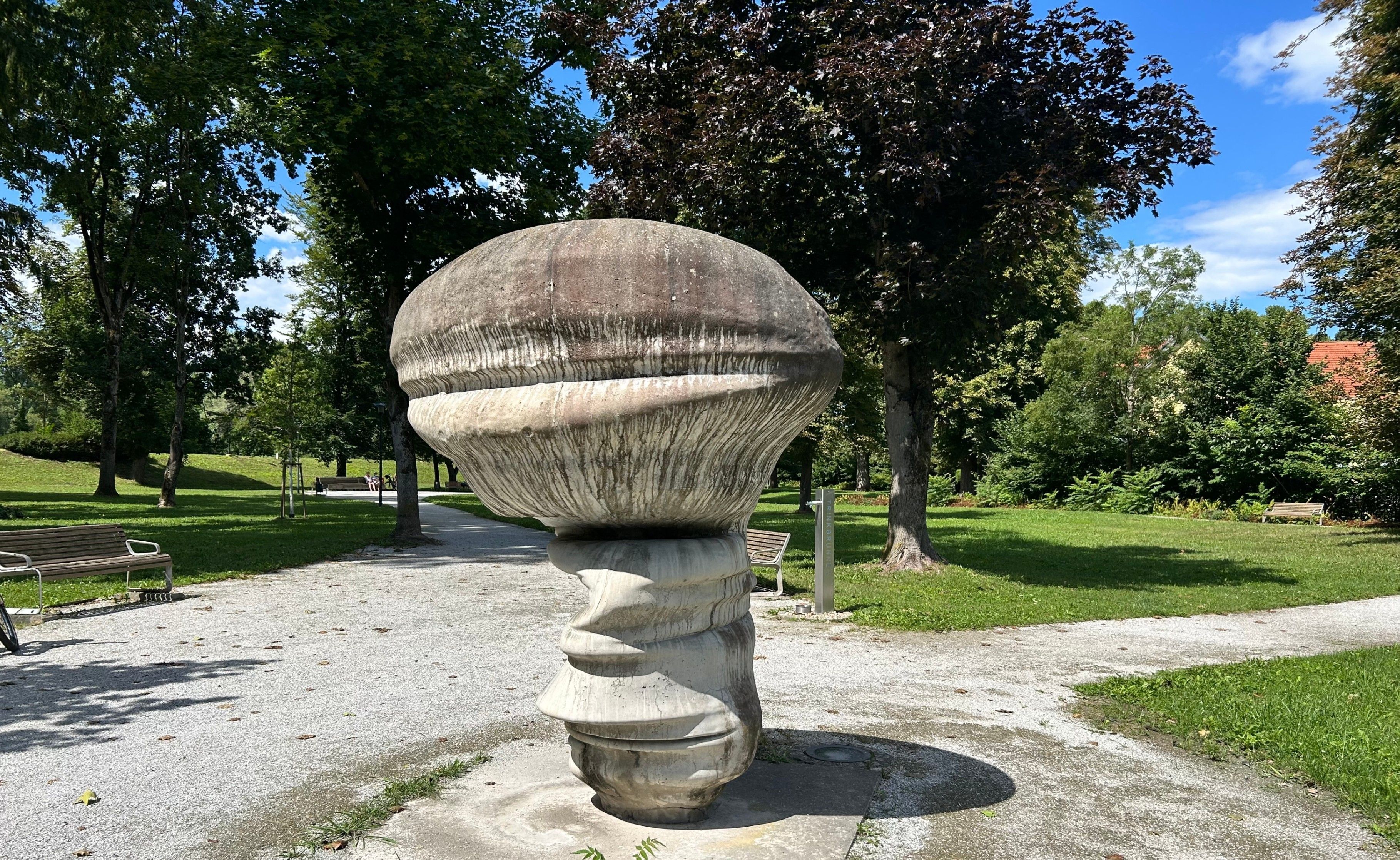 Eine große Steinskulptur in Form eines Pilzes steht in einem Park mit klarem blauem Himmel und üppiger Grünfläche im Hintergrund.
