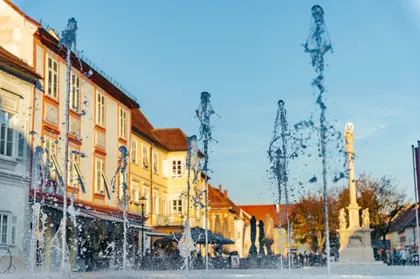 Ein Stadtplatz mit mehreren Wasserbrunnen vor einem Gebäude mit vielen Fenstern. Im Hintergrund ist eine Statue zu sehen.
