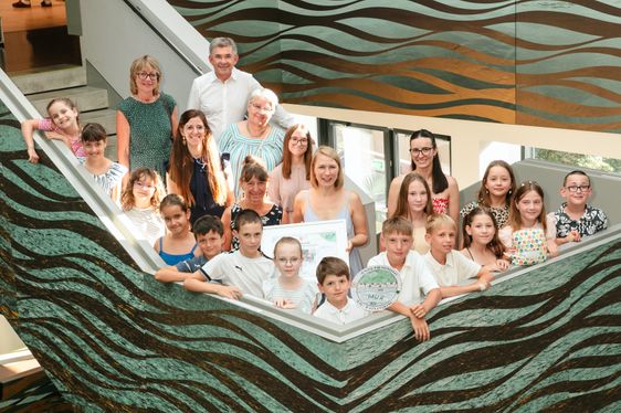 Eine Gruppe von Menschen, Erwachsene und Kinder, posiert für ein Foto und hält ein Zertifikat. Sie stehen in einem Raum mit einem wellenförmigen Wanddesign.