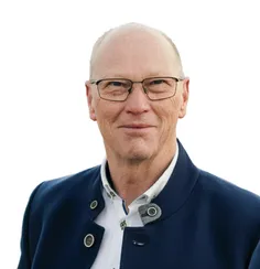 Ein älterer Mann mit Brille, lächelnd, trägt einen dunkelblauen Blazer mit zwei Knöpfen links und einem rechts sowie ein weißes Hemd.