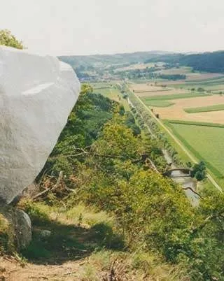 Ein Panoramablick von einem Hügel zeigt einen großen Felsen, Grünflächen und einen Fluss mit einer Brücke. Die Landschaft ist gefüllt mit grünen Feldern, Bäumen und einem fernen Horizont.