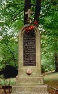 Ein Grabstein steht in einem Wald, geschmückt mit einem Kreuz, Blumen und Blättern. Der Grabstein hat eine gravierte Inschrift mit Namen und Daten.