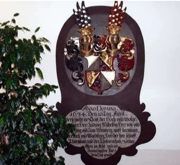 An der weißen Wand hängt eine Holztafel mit einem Wappen in goldenen Farben und einer lateinischen Inschrift. Die Tafel ist von dekorativen Elementen umgeben. Links neben der Tafel steht eine grüne Pflanze.