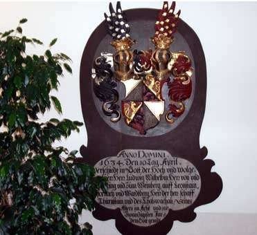 An der weißen Wand hängt eine Holztafel mit einem Wappen in goldenen Farben und einer lateinischen Inschrift. Die Tafel ist von dekorativen Elementen umgeben. Links neben der Tafel steht eine grüne Pflanze.