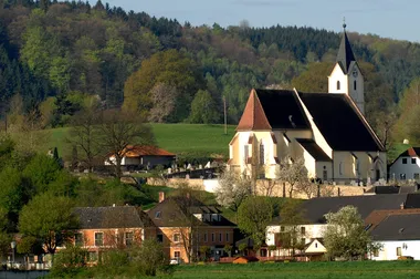 Eine Kirche steht prominent in einem üppigen grünen Tal, umgeben von Häusern und einem Friedhof. Die Landschaft wird von dichten Bäumen und einem klaren Himmel eingerahmt.