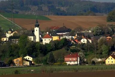 Ein malerisches Dorf mit verschiedenen Häusern, einer Kirche mit einem Turm und einer umliegenden Landschaft aus Feldern und Grün unter einem klaren Himmel.