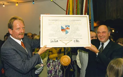 Ein offizielles Dokument wird von zwei Männern vor einer Menge gehalten. Es scheint sich um ein Zertifikat oder Diplom aus dem Niederösterreichischen Landesarchiv zu handeln. Das Dokument zeigt ein Siegel und Unterschriften.