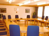Ein Konferenzraum mit einem langen Holztisch, blauen Stühlen und weißen Wänden. Es gibt Bilderrahmen, eine Topfpflanze und Blumen auf dem Tisch. Der Raum hat Deckenleuchten und Fenster mit Vorhängen.