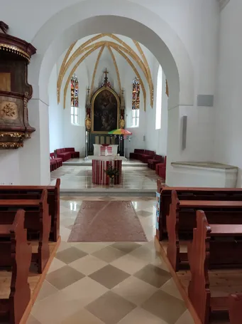 Das Innere einer Kirche zeigt einen Hauptaltar, der mit einem Gemälde geschmückt ist, flankiert von Buntglasfenstern und goldenen Statuen. Rote Stühle sind in Reihen angeordnet, und ein bunter Regenschirm ruht in der Mitte. Der Boden ist kariert und ein Teppich liegt davor.