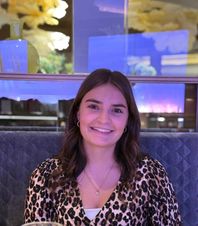 Eine Frau mit langen braunen Haaren und einem Leopardenmuster-Oberteil sitzt in einer Restaurant-Loge und lächelt für ein Foto.