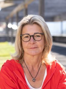 Eine Frau mit blondem Haar, die eine Brille und eine rote Jacke trägt, lächelt und schaut in die Kamera.