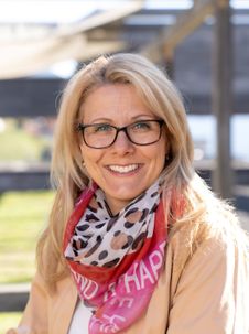 Eine Frau mit blondem Haar, die eine Brille und ein Halstuch mit dem Wort 'Glücklich' trägt, lächelt und posiert für ein Foto.