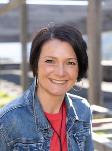 Eine Frau mit kurzen schwarzen Haaren lächelt und trägt eine Jeansjacke und ein rotes Shirt.