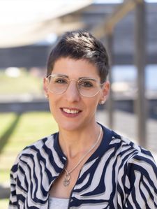 Eine Frau mit kurzen Haaren und einer Brille lächelt und schaut in die Kamera. Sie trägt eine Zebra-Print-Jacke und eine Halskette.