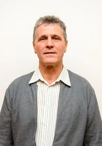 Ein Mann mit grauen Haaren und einer grauen Strickjacke steht vor einer weißen Wand. Er trägt ein weißes Hemd mit grünen Streifen.