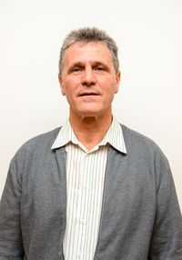 Ein Mann mit grauen Haaren und einer grauen Strickjacke steht vor einer weißen Wand. Er trägt ein weißes Hemd mit grünen Streifen.