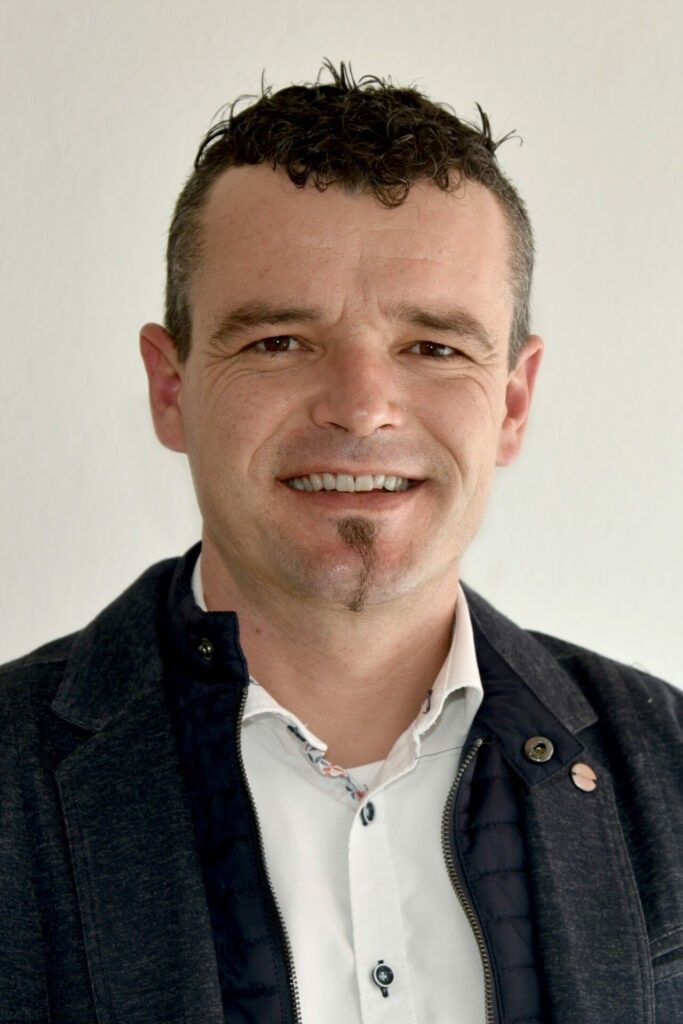 Ein Mann mit lockigem Haar, einem Schnurrbart und einem Bart lächelt und schaut in die Kamera. Er trägt ein weißes Hemd und eine schwarze Jacke.