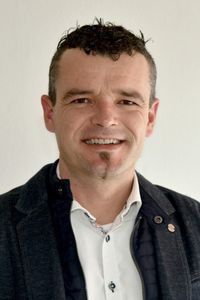 Ein Mann mit lockigem Haar, einem Schnurrbart und einem Bart lächelt und schaut in die Kamera. Er trägt ein weißes Hemd und eine schwarze Jacke.