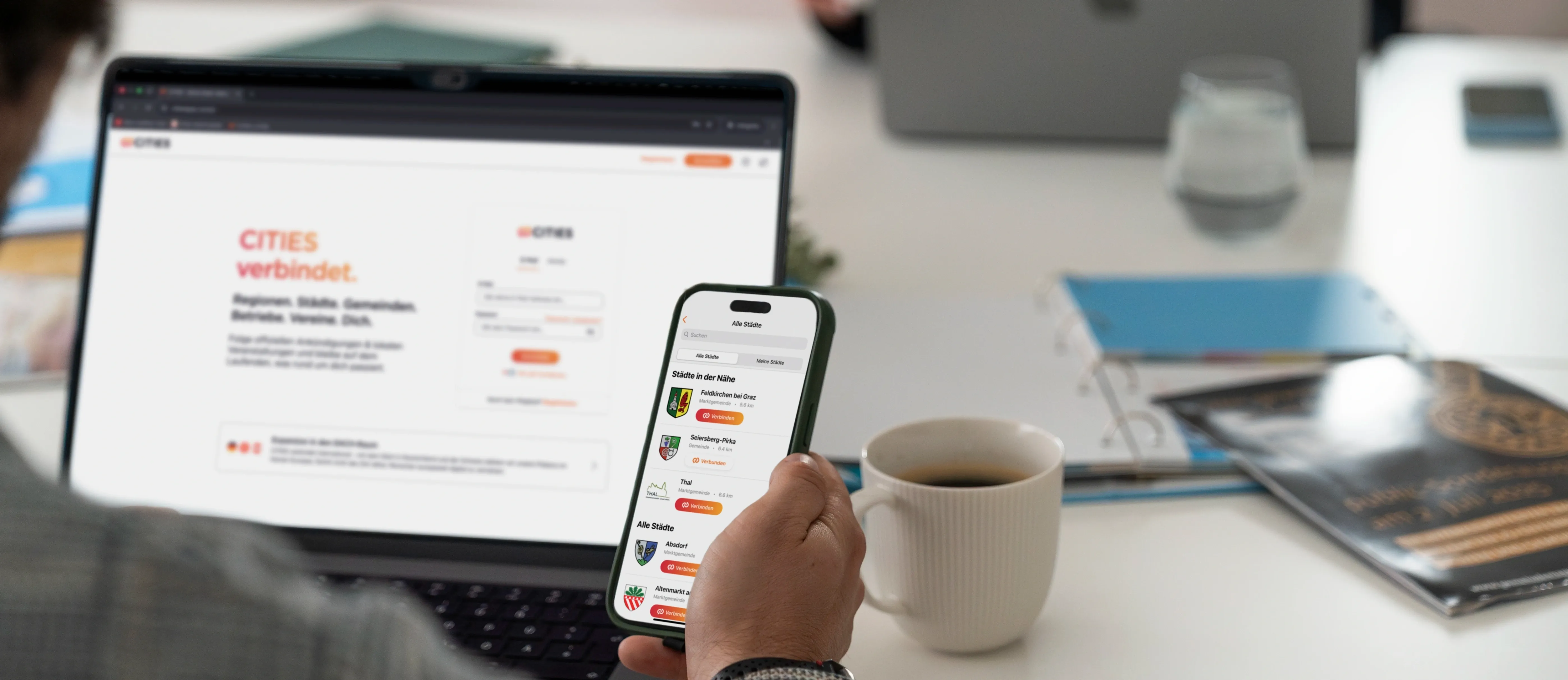 Eine Person benutzt ein Smartphone, das eine Karten-App mit nahegelegenen Städten anzeigt. Der Bildschirm zeigt eine Liste von Städten mit Entfernungen und Symbolen. Die Person sitzt an einem Schreibtisch mit einem Laptop und einer Tasse Kaffee.