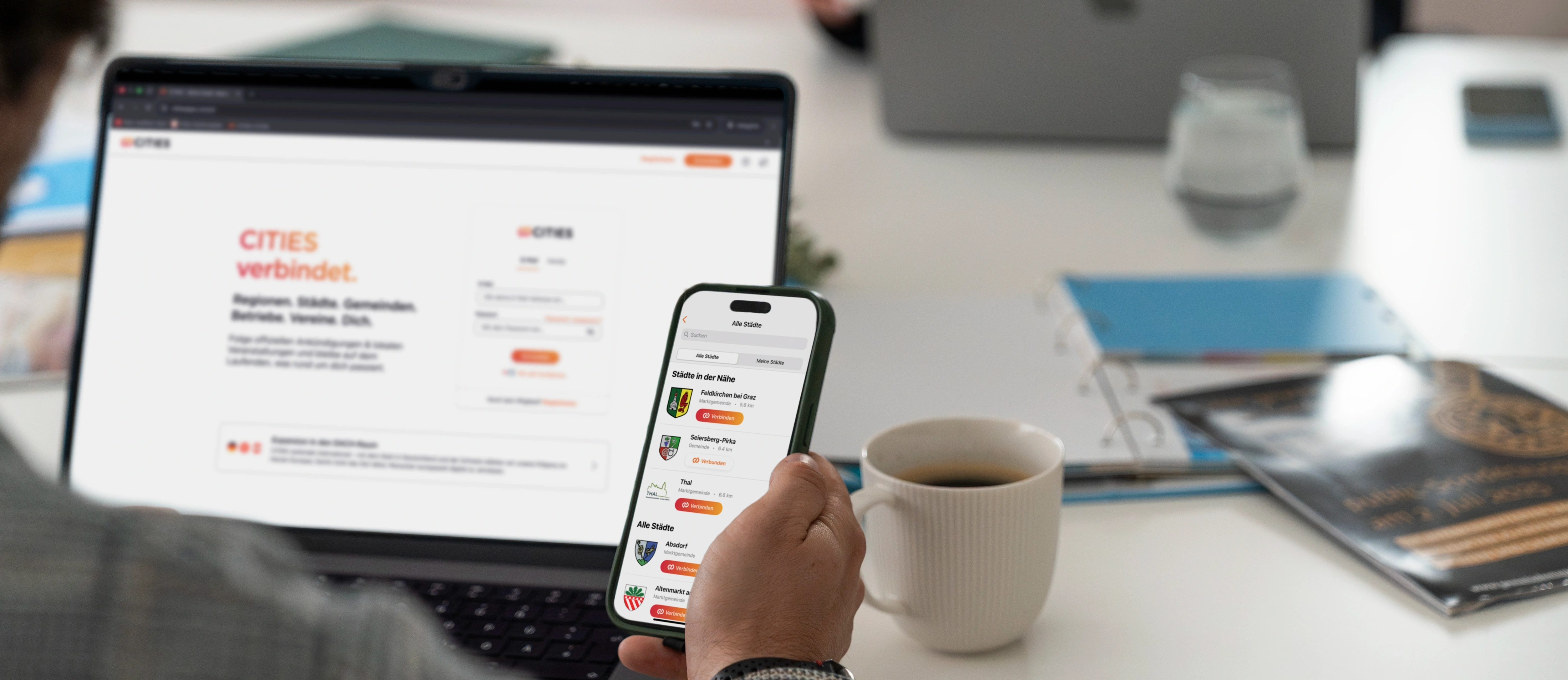 Eine Person benutzt ein Smartphone, das eine Karten-App mit nahegelegenen Städten anzeigt. Der Bildschirm zeigt eine Liste von Städten mit Entfernungen und Symbolen. Die Person sitzt an einem Schreibtisch mit einem Laptop und einer Tasse Kaffee.