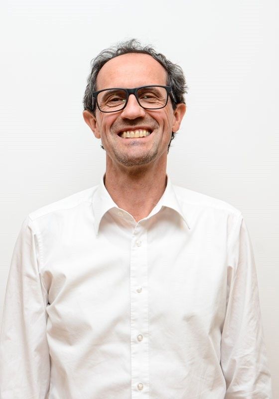 Ein Mann mit grauem Haar, der eine Brille und ein weißes Hemd trägt, lächelt und posiert für ein Foto.
