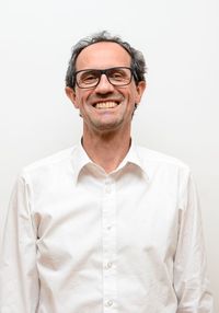 Ein Mann mit grauem Haar, der eine Brille und ein weißes Hemd trägt, lächelt und posiert für ein Foto.