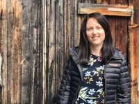 Eine Frau mit dunklem Haar lächelt vor einer Holzwand. Sie trägt eine schwarze Daunenjacke über einem Blumenhemd.