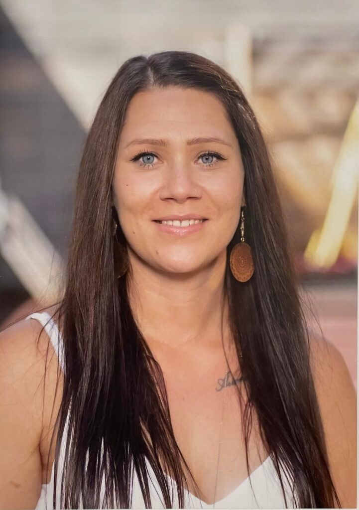 Eine Frau mit langen dunklen Haaren und blauen Augen lächelt in die Kamera. Sie trägt ein weißes ärmelloses Oberteil und hat eine Tätowierung auf ihrer Brust.